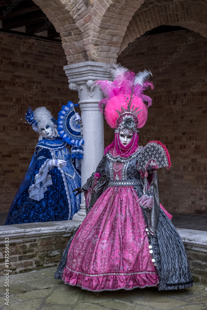 Masked women in ornate blue and pink costumes with fans in an inner courtyard during the Venice Carnival (Carnivale di Venezia)