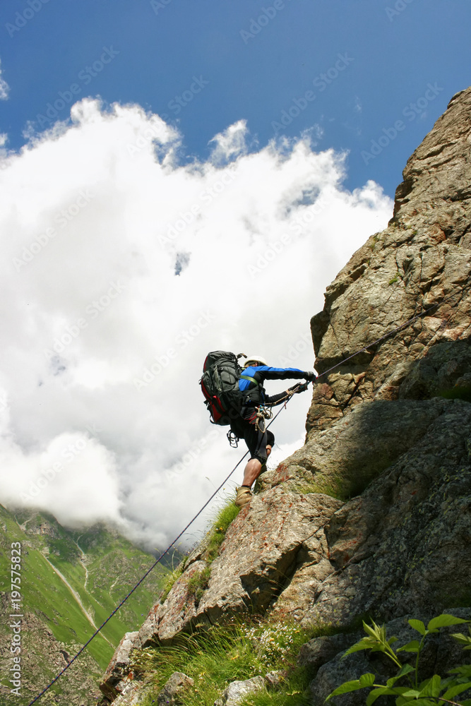 The climber climbs the rock by the rope. Big backpack, helmet, various equipment. Sunny day, mountain valley with alpine meadows and flowers. In the background there is a blue sky with light clouds.
