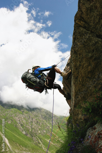 The climber climbs the rock by the rope. Big backpack, helmet, various equipment. Sunny day, mountain valley with alpine meadows and flowers. In the background there is a blue sky with light clouds.