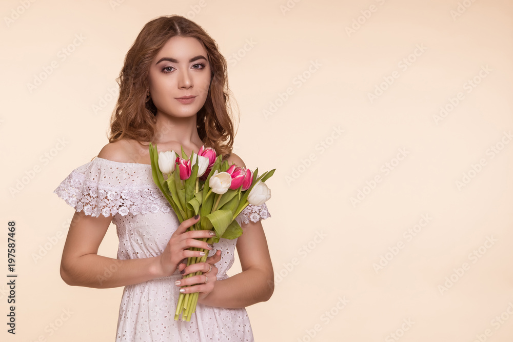 Beautiful young woman stands on a beige background, in a white dress and a bouquet of tulips. International Women's Day, March 8. Copy-space. Beautiful young woman stands on a beige background, in a white dress and a bouquet of tulips. International Women's Day, March 8. Copy-space.