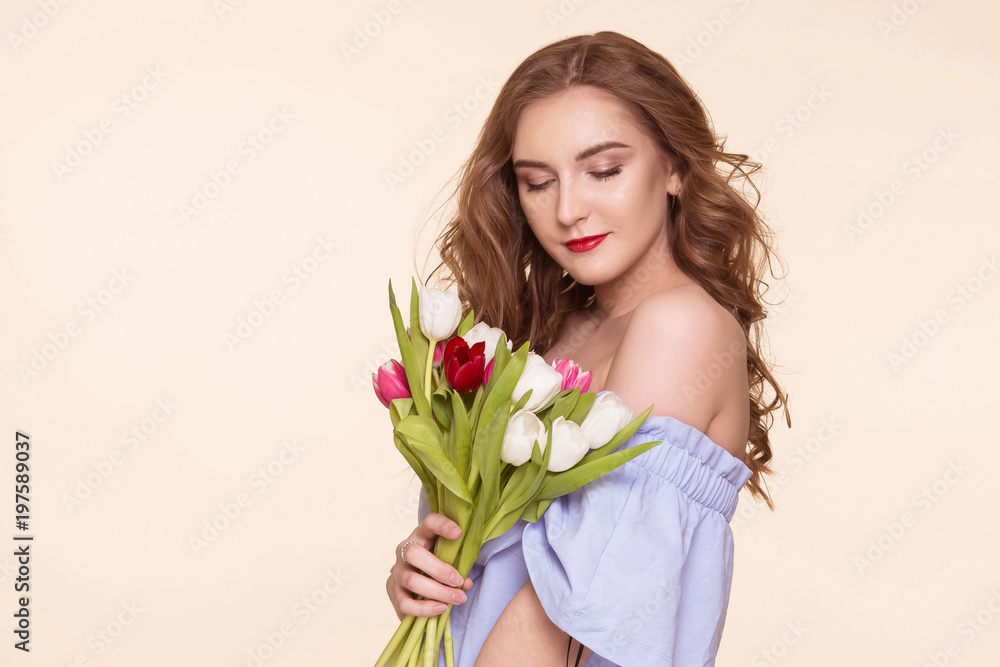 Beautiful young girl with a tulip bouquet standing on a beige, background, in a blue dress. Women's Day, March 8th. Copy-space.