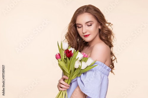 Beautiful young girl with a tulip bouquet standing on a beige, background, in a blue dress. Women's Day, March 8th. Copy-space.