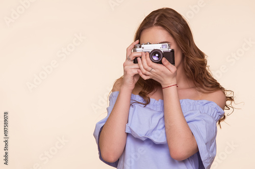 A beautiful young girl in a pin-up image takes pictures on an old DSLR, on a beige background. Metal camera.