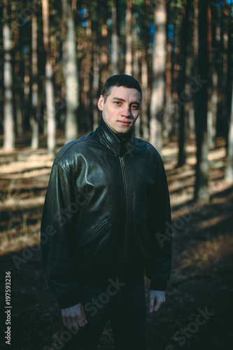 A young man of criminal appearance in a black leather jacket posing in an autumn forest.