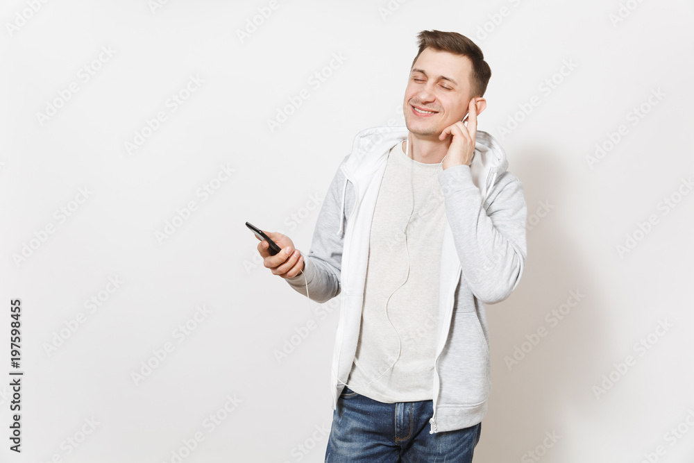 Young handsome smiling man student in t-shirt, blue jeans and light sweatshirt listening to music on earphones on mobile phone and enjoys in studio on white background. Concept of emotions, good mood. Young handsome smiling man student in t-shirt, blue jeans and light sweatshirt listening to music on earphones on mobile phone and enjoys in studio on white background. Concept of emotions, good mood.