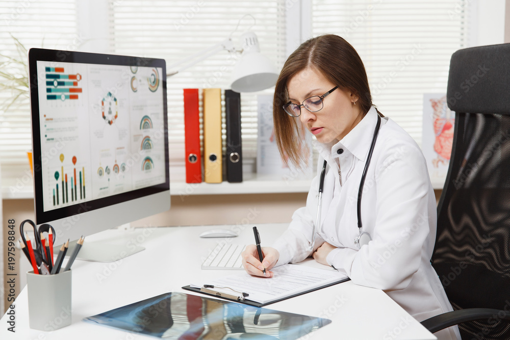 Young woman sitting at desk, working on computer, filling out medical documents in light office in hospital. Female doctor in medical gown, stethoscope in consulting room. Healthcare, medicine concept Young woman sitting at desk, working on computer, filling out medical documents in light office in hospital. Female doctor in medical gown, stethoscope in consulting room. Healthcare, medicine concept