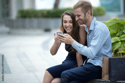 Young couple using smartphone together, paper shopping bags near them