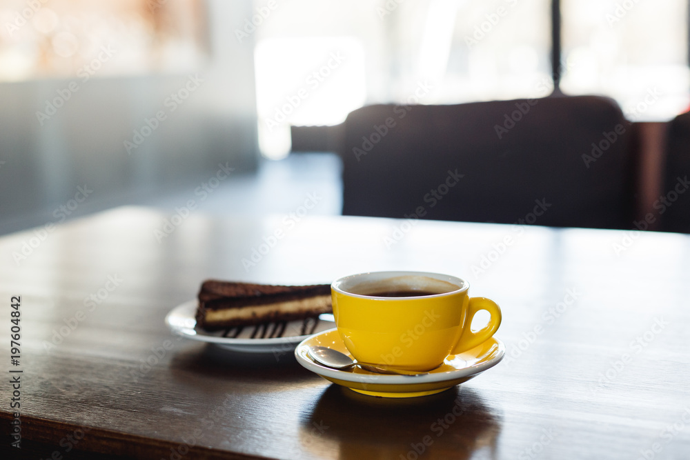 A horizontal image of a yellow cup of black coffee and a plate with a piece of chocolate cake. Bokeh background. A horizontal image of a yellow cup of black coffee and a plate with a piece of chocolate cake. Bokeh background.
