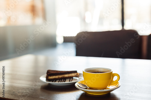 A horizontal image of a yellow cup of black coffee and a plate with a piece of chocolate cake. Bokeh background.