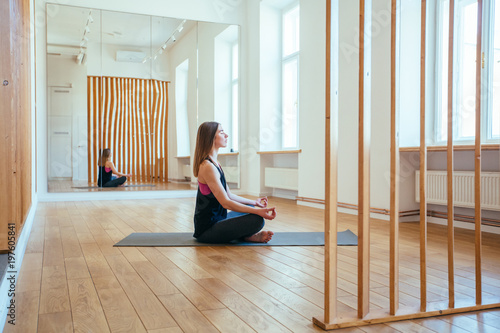 Woman pracitcing yoga sitting in empty room with mirror. Female relaxing in Padmasana exercise, Lotus pose, mudra gesture, full length, middle aged slim woman training at home, side view photo
