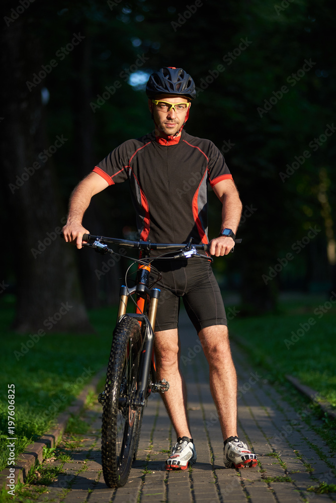 Obraz premium Sporty smiling man bicyclist walking near bicycle happy to begin morning with training. Sportsman having fun exercising outdoors. Concept of healthy lifestyle, new beginning