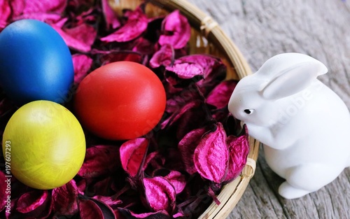 Happy Easter Day! White rabbit and colors Easter eggs in basket with pink flower decoration on wooden table background top view