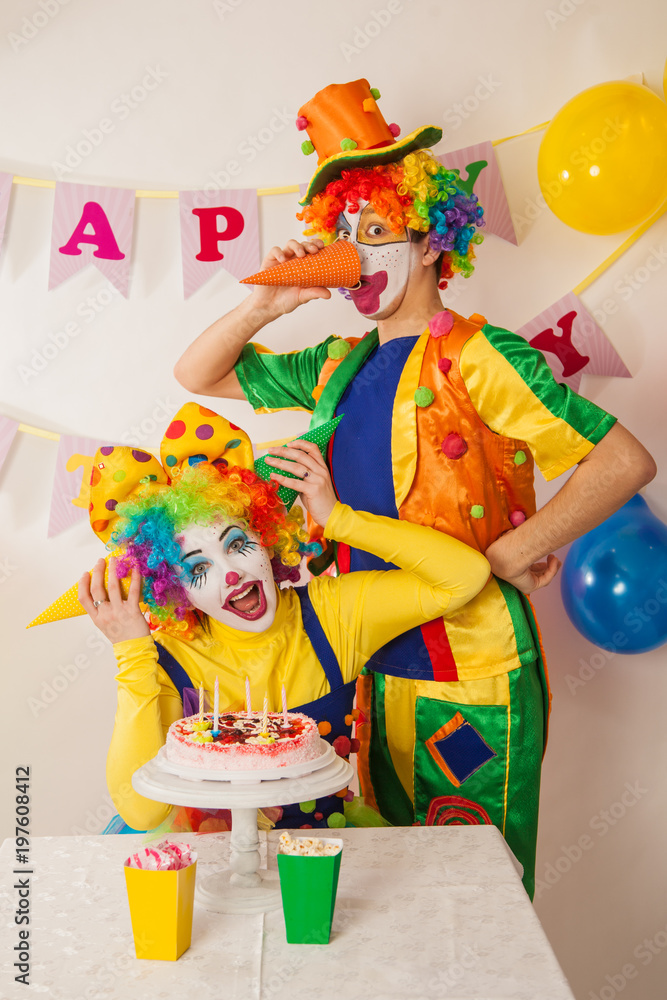 Clowns are a boy and a girl in bright costumes at the child's birthday. A table with refreshments and a cake. The explosion of emotions and the fun of the circus. greed and desire to eat cake Clowns are a boy and a girl in bright costumes at the child's birthday. A table with refreshments and a cake. The explosion of emotions and the fun of the circus. greed and desire to eat cake
