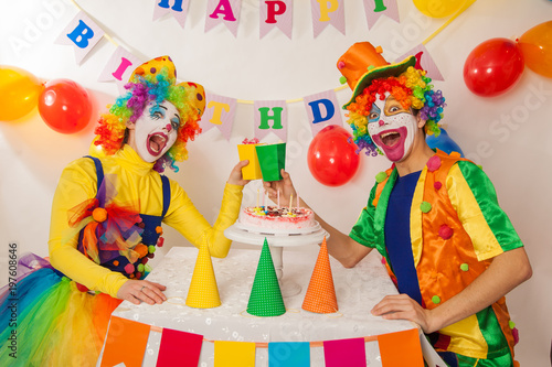Clowns are a boy and a girl in bright costumes at the child's birthday. A table with refreshments and a cake. The explosion of emotions and the fun of the circus. greed and desire to eat cake © izida1991