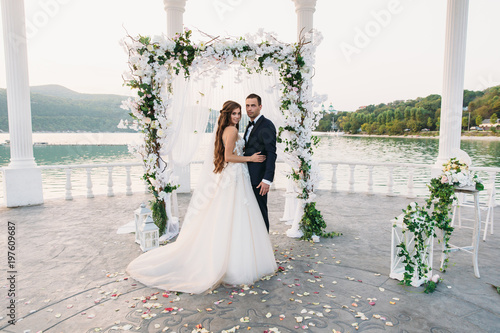 Attractive groom and bride at wedding day ceremory with arch and lake on background stands together. Beautiful newlyweds, young woman in white dress and long hairs, men in black suit. Happy family