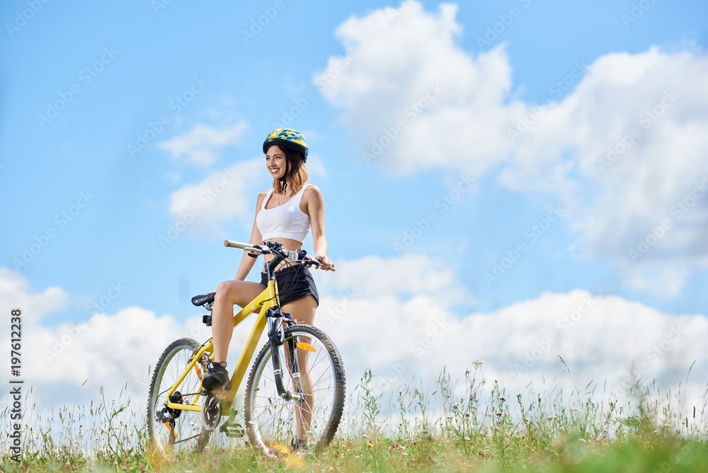 Happy young female rider cycling on yellow bicycle on a grass, wearing helmet, enjoying sunny day in the mountains against blue sky with clouds. Outdoor sport activity, lifestyle concept. Copy space