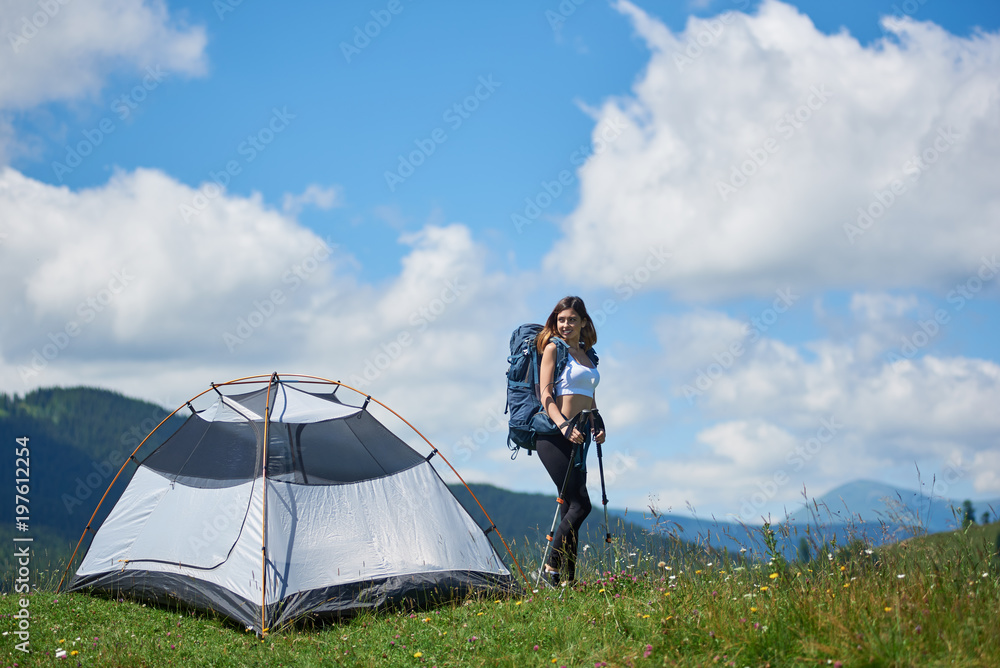 Attractive girl tourist with backpack and trekking sticks near tent on the top of a hill against blue sky and clouds, smiling, looking away, enjoying summer morning in the mountains.