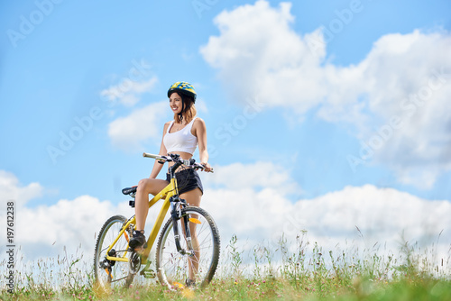 Happy young female rider cycling on yellow bicycle on a grass, wearing helmet, enjoying sunny day in the mountains against blue sky with clouds. Outdoor sport activity, lifestyle concept. Copy space © anatoliy_gleb