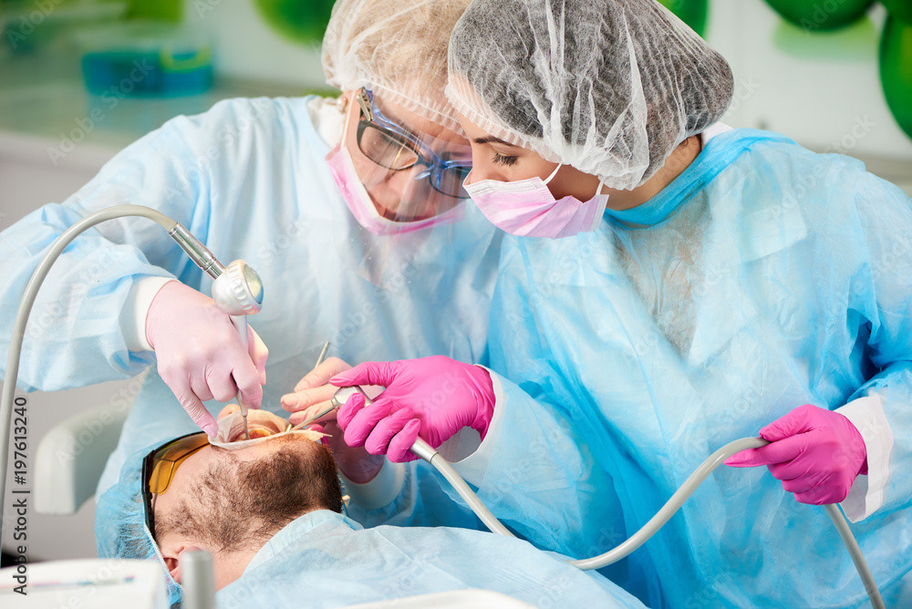 Two dentists in blue robes are scaling and polishing the teeth of a male client of a dental