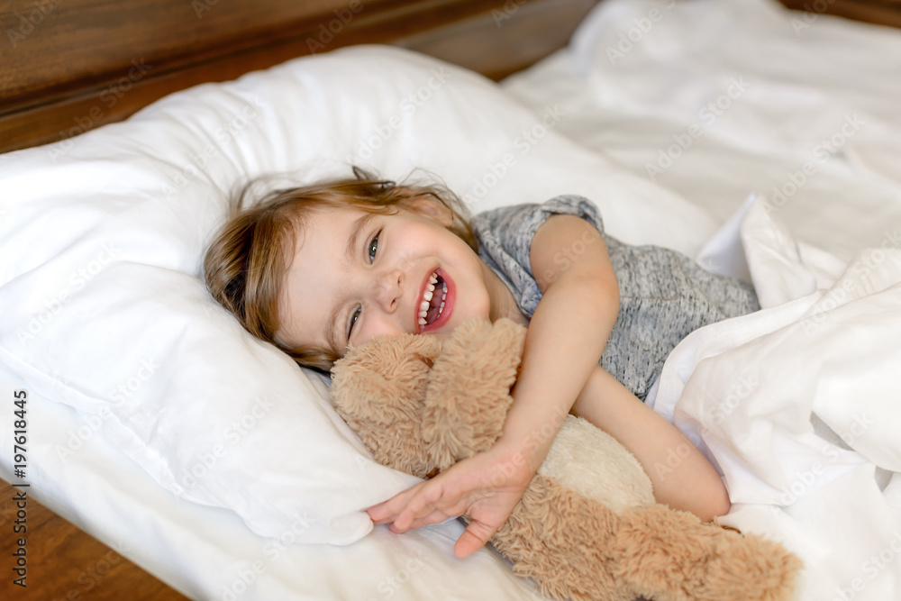 Active adorable baby wakes up lying in bed with her Teddy bear toy, soft white linens. Children's day. World Smile Day. Start the morning right! Start the morning with a smile! Active adorable baby wakes up lying in bed with her Teddy bear toy, soft white linens. Children's day. World Smile Day. Start the morning right! Start the morning with a smile!