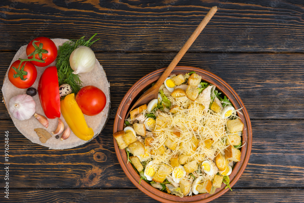 High angle view of large clay dish with tasty caesar salad next to vegetables on wooden desk