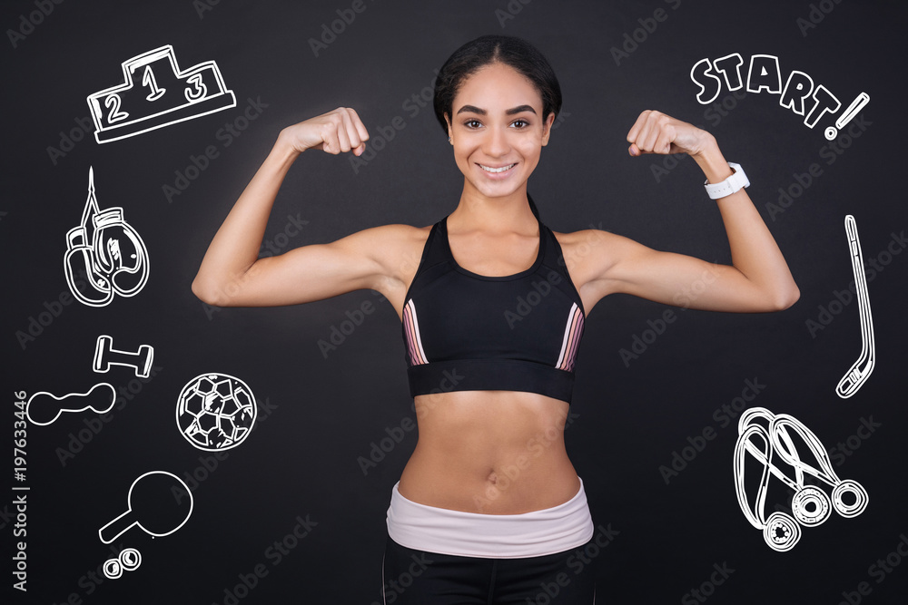 Strong sportswoman. Cheerful active sporty young woman looking happy and feeling proud of her strength while showing biceps and smiling