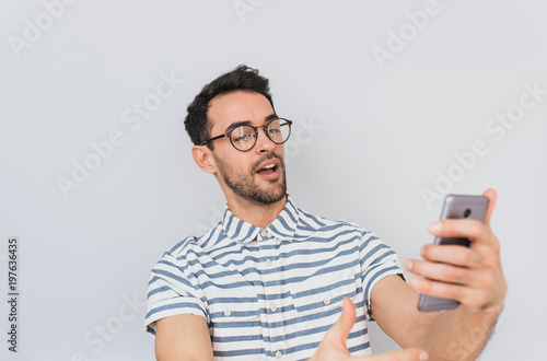 Happy young European bearded man wears striped shirt and round glasses, making selfie and browsing newsfeed via social networks using smart phone, looking at screen with cheerful smile. People concept © iuricazac