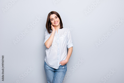 Portrait of trendy, cheerful, modern girl in denim outfit, holding hands in pocket of jeans and touching her neck, looking at camera, having short hair, isolated on grey background