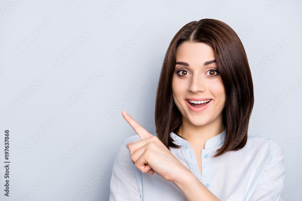 Close up portrait with copy space of charming, cheerful, pretty,  brunette, nice, stylish, trendy woman inshirt, pointing  forefinger on empty place, looking at camera, isolated on grey background