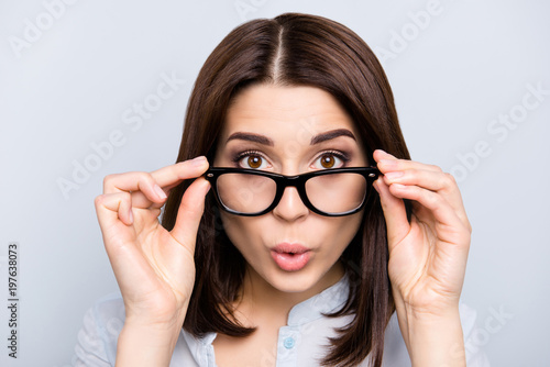 Close up portrait of shocked, impressed, pretty, charming, cute, successful, professional girl looking out glasses, holding eyelets of spectacles on face with two hands, isolated on grey background