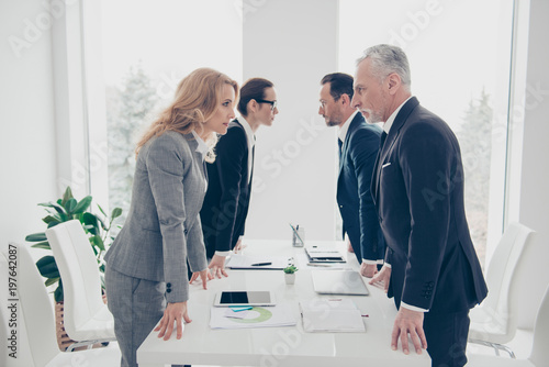 Business competition, four stylish business persons in suits having disagreement and conflict, standing together near desktop in front of each other, face to face with disrespect expression photo