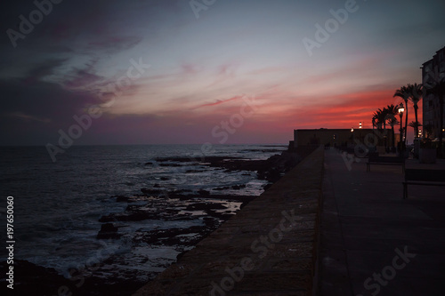 Incredible red-pink sunset in Cadiz, Spain. City promenade with palm trees and lanterns.