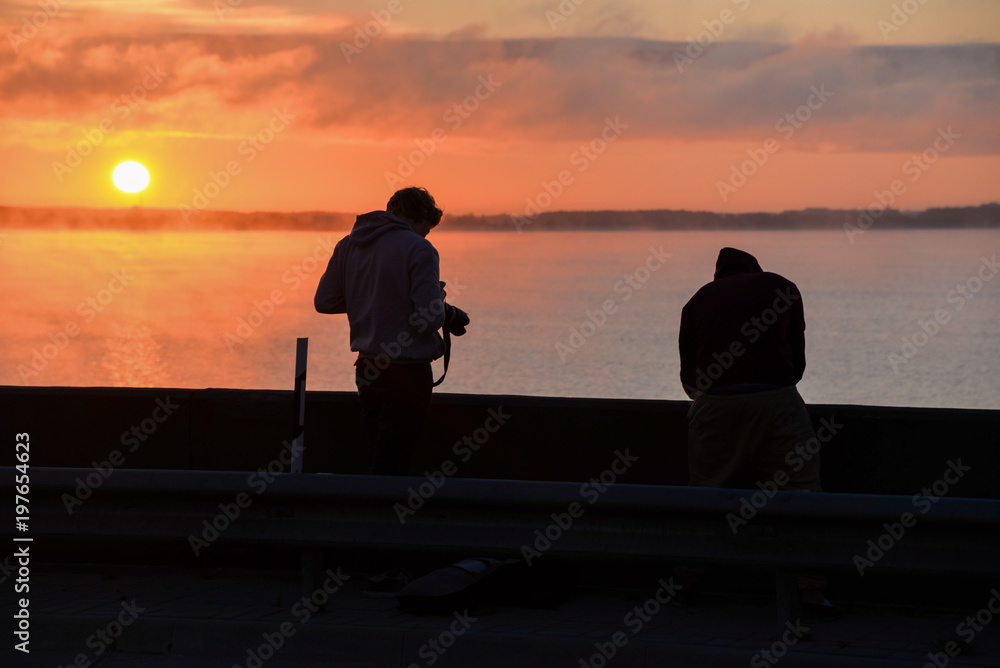 Two young filmmakers filming an early sunrise with an orange sky and sun Two young filmmakers filming an early sunrise with an orange sky and sun