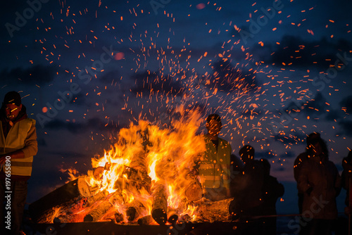 Large burning bonfire with soft glowing flame and sparkles flying all around. Romantic summer evening, people relaxing and enjoying calmness at the seaside during the Night of ancient lights.