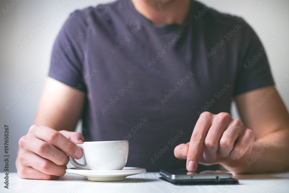 Young man drinking morning coffee and holding mobile phone. Coffee break. Man holding cup of fresh roasted coffee and looking at the smart phone. Young man drinking morning coffee and holding mobile phone. Coffee break. Man holding cup of fresh roasted coffee and looking at the smart phone.