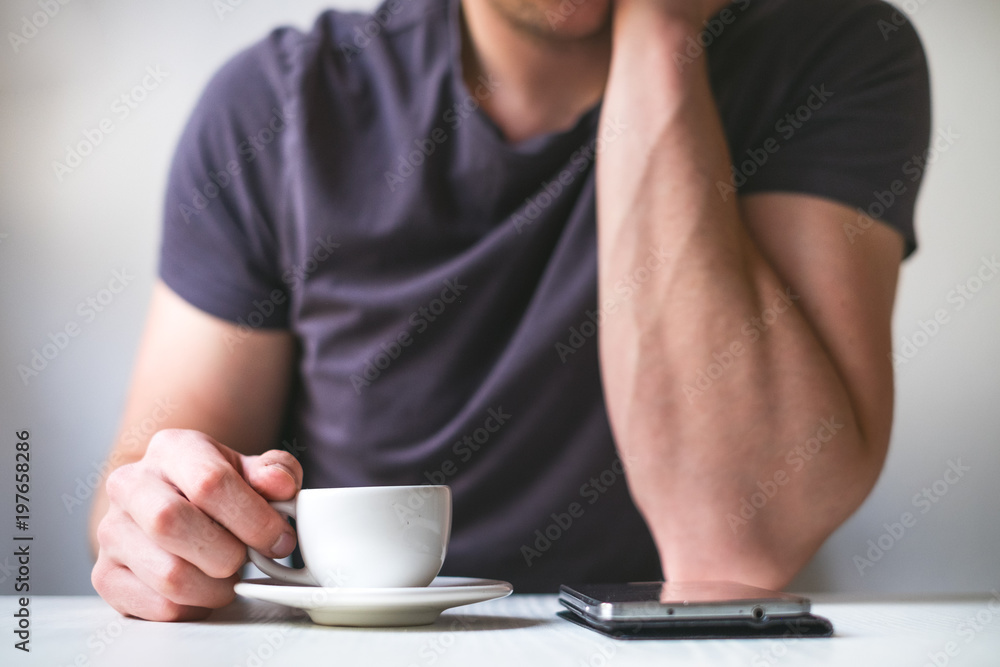 Young man drinking morning coffee and holding mobile phone. Coffee break. Man holding cup of fresh roasted coffee and looking at the smart phone. Young man drinking morning coffee and holding mobile phone. Coffee break. Man holding cup of fresh roasted coffee and looking at the smart phone.