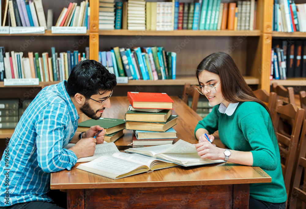Two smart students working on home assignment, reading books attentively, sitting at square library table next to the bookshelves Two smart students working on home assignment, reading books attentively, sitting at square library table next to the bookshelves