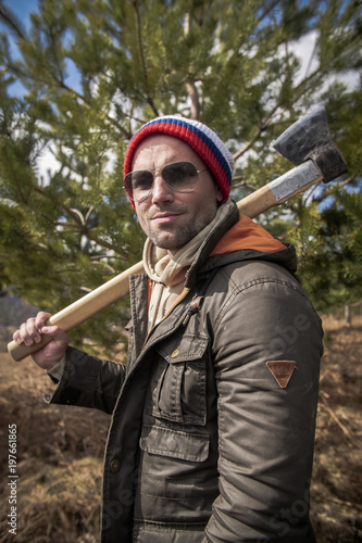 One handsome strong stylish male lumberjack holding wooden axe in forest outdoor and mountains in the background