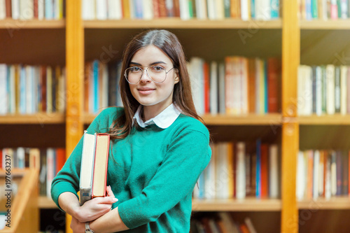 Portrait of young beautiful student girl wears glasses and green sweater holding books before bookshelves in the library