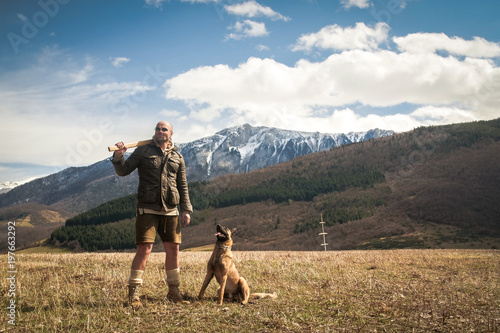 Handsome Man Lumberjack and his Belgian Shepherd Posing in Front of a Camera With Mountains In The Background