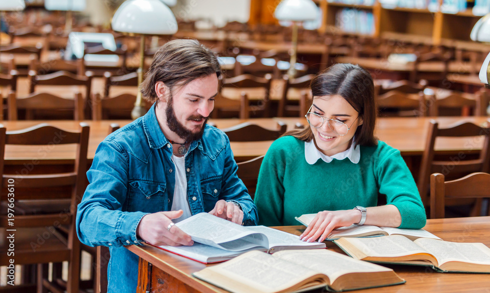 Two smart students working on home assignment, reading books attentively, sitting at square library table in the reading hall Two smart students working on home assignment, reading books attentively, sitting at square library table in the reading hall