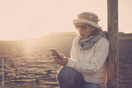 woman using phone outdoor sitting down ant the beach with beautiful sunset in the background