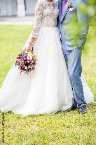 Bride and Groom Standing Hugging Together in Grass with Purple and Pink Bouquet of Flowers