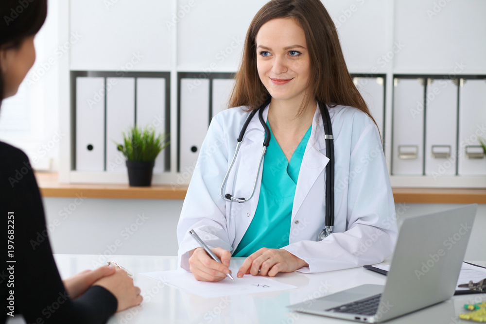 Beautiful female doctor and patient having a talk while sitting at the desk. Physician writes prescription with silver pen at workplace in office. Medicine, pharmacology, prescribing treatment and Beautiful female doctor and patient having a talk while sitting at the desk. Physician writes prescription with silver pen at workplace in office. Medicine, pharmacology, prescribing treatment and