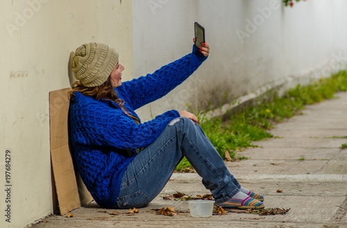 Outdoor view of homeless woman begging on the street in cold autumn weather sitting on the floor at sidewalk taking a selfie photo