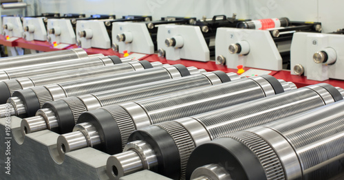 Lined up magnetic cylinders for die cut on rotary printing press. Magnetic cylinder for flexo rotary die cutting. Magnetic roll and in-line press machine in background. Cylinder for cutting dies.