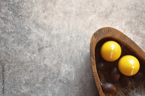 Top view of five brown chocolate easter eggs and two yellow candles in wooden plate on grunge grey background with space for text. Happy Easter still life.