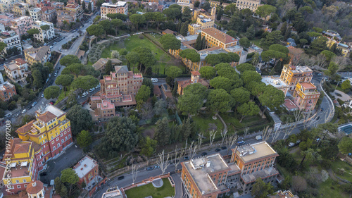 Aerial view of a small square with trees and meadows on either side of a road. The street passes under an ancient Rome arch.