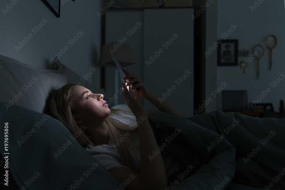 Sleepy exhausted woman lying in bed using smartphone at late night, can not sleep. Insomnia, nomophobia, sleep disorder concept. Addiction on social networks - obrazy, fototapety, plakaty 