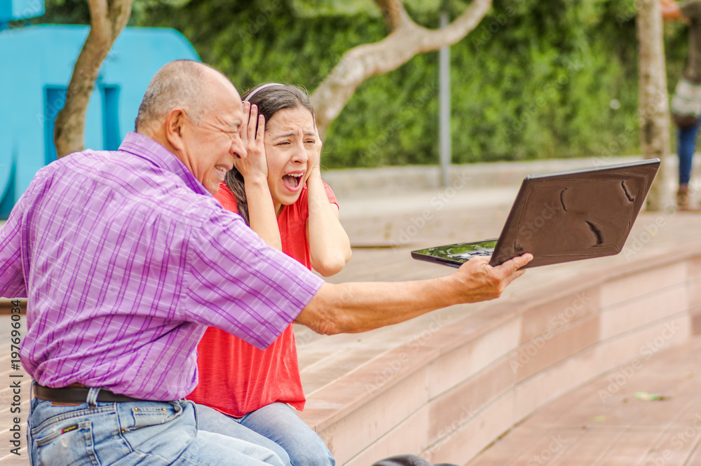 Outdoor view of father holding and computer and sacry daugher screaming afraid of his dad throw the computer in the ground, at park Outdoor view of father holding and computer and sacry daugher screaming afraid of his dad throw the computer in the ground, at park